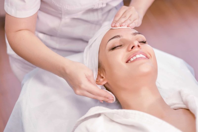 An esthetician cleansing a smiling client's face during a facial